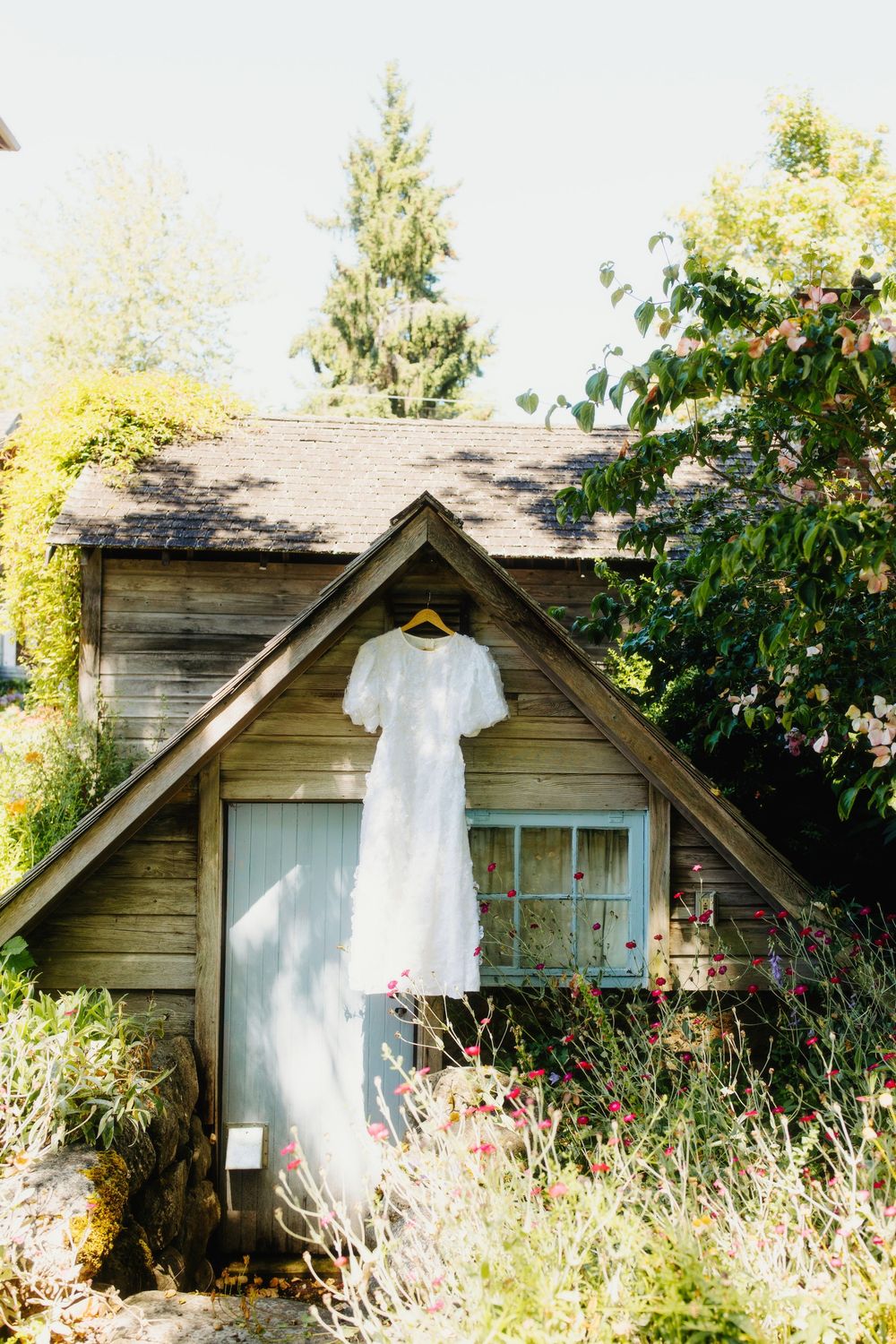White wedding dress hanging outside a rustic wooden cottage surrounded by wildflowers.