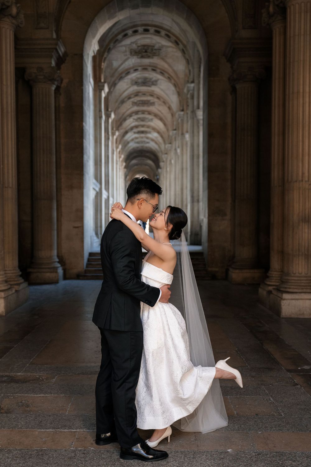 A romantic wedding couple shares intimate moments in a grand arched hallway with classical architecture.