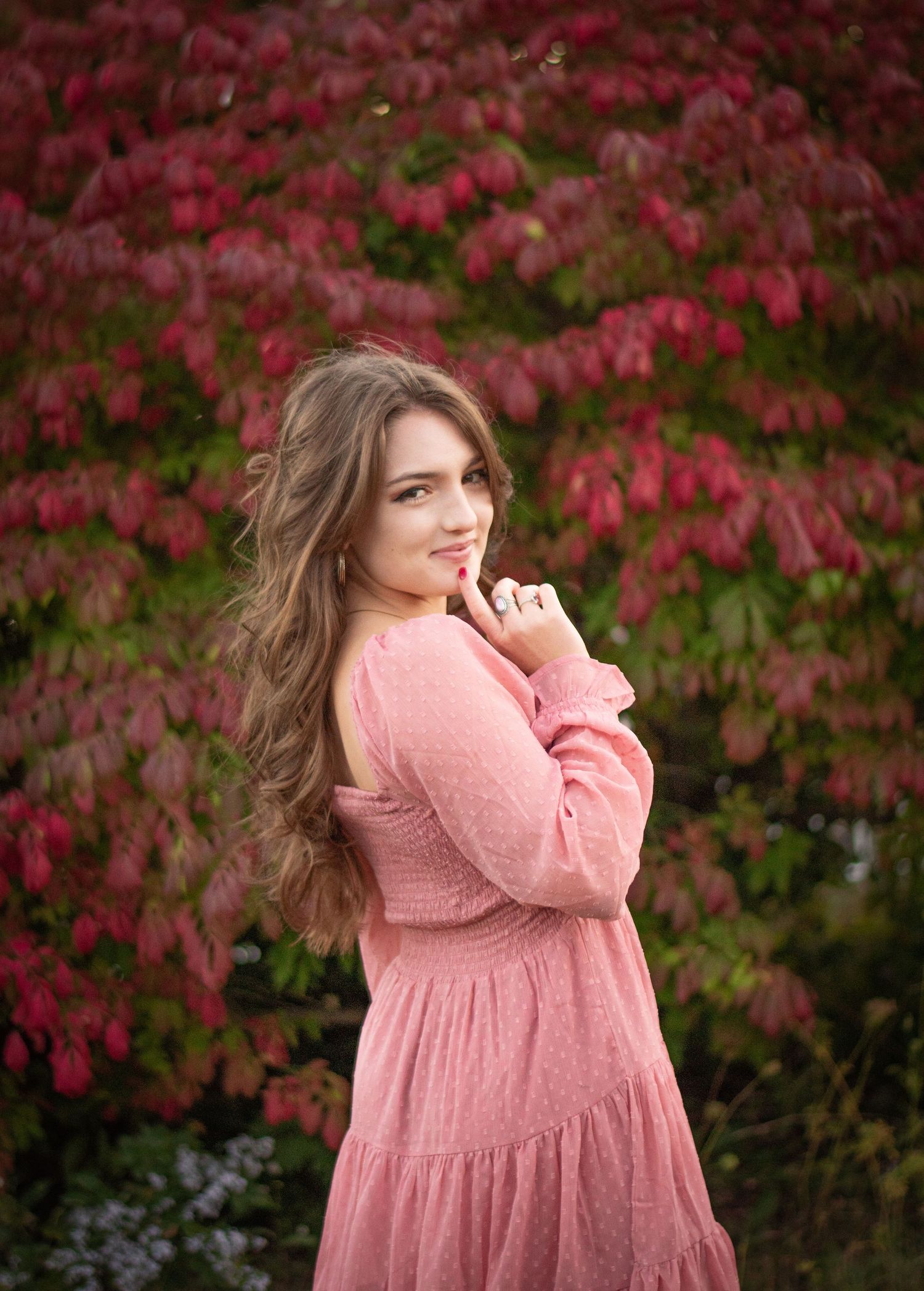 Senior Teen girl with wavy hair in pink sweater leaning on white picket fence outdoors. Photo by Xenophon Photography in Maine.