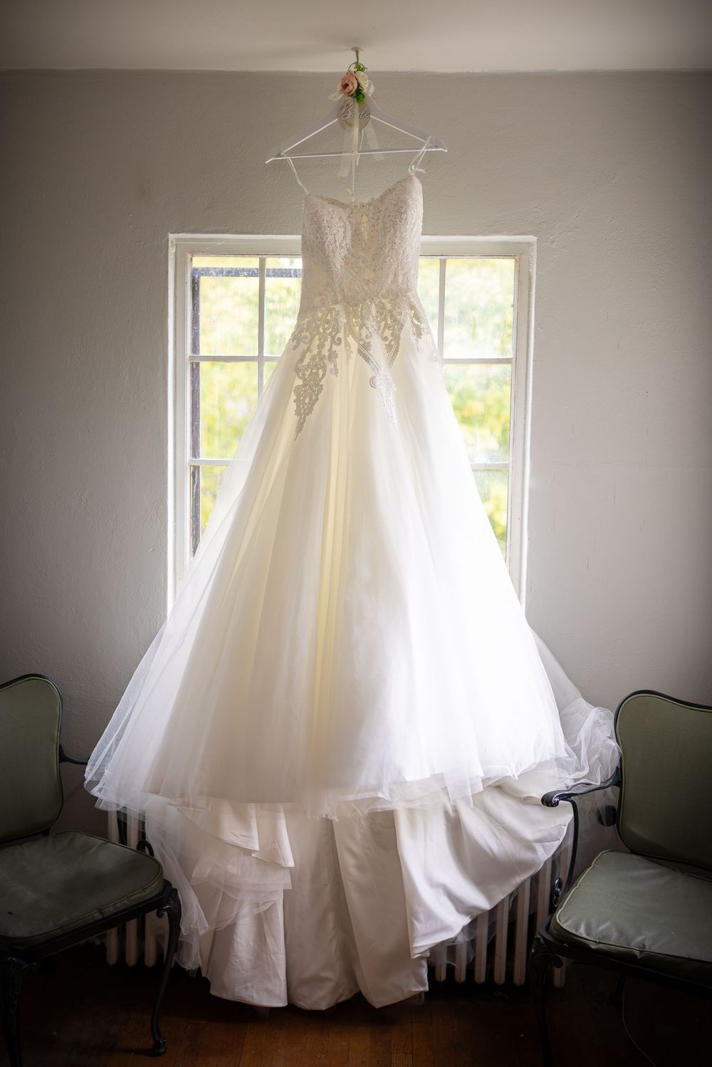 A white wedding dress with lace details hangs elegantly by a window in a bright room.