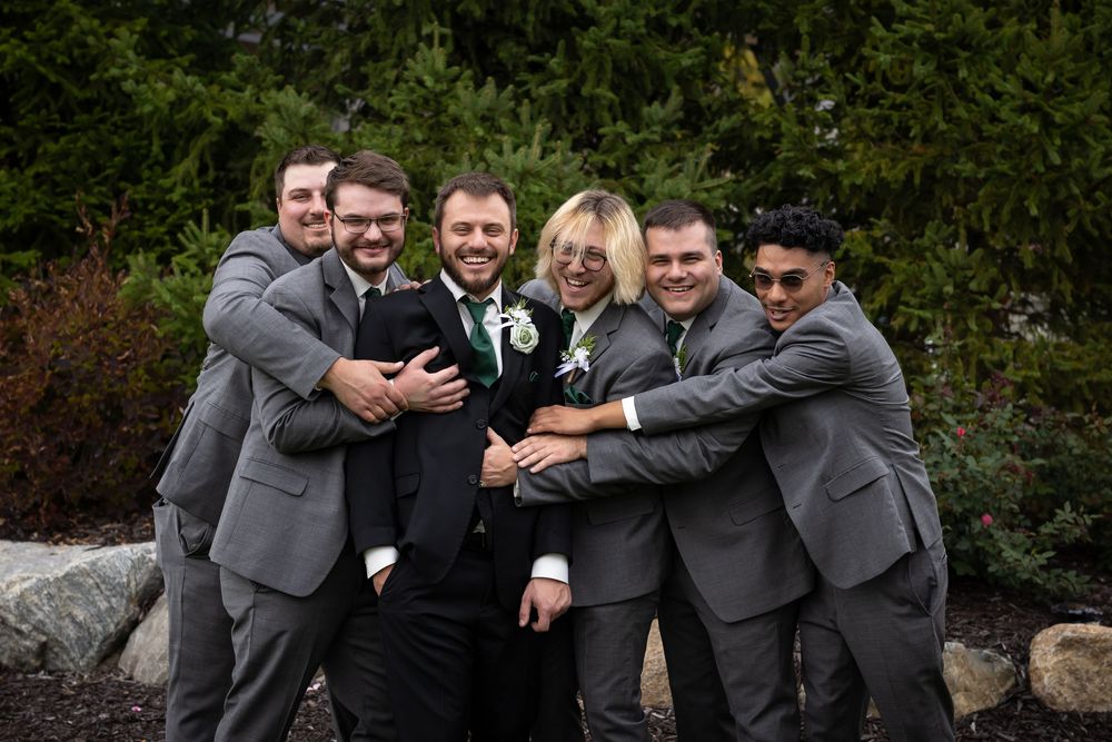 Groomsmen in gray suits share a playful group hug outdoors during a wedding celebration.