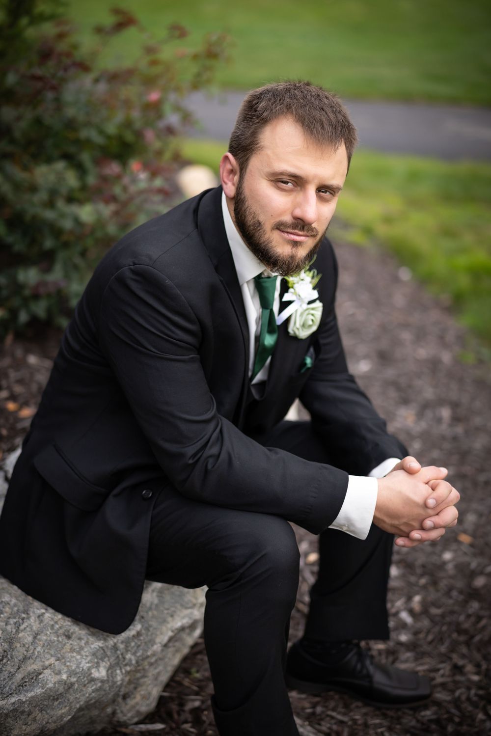 A groom in a black tuxedo sits thoughtfully on a stone wall in an outdoor garden setting.