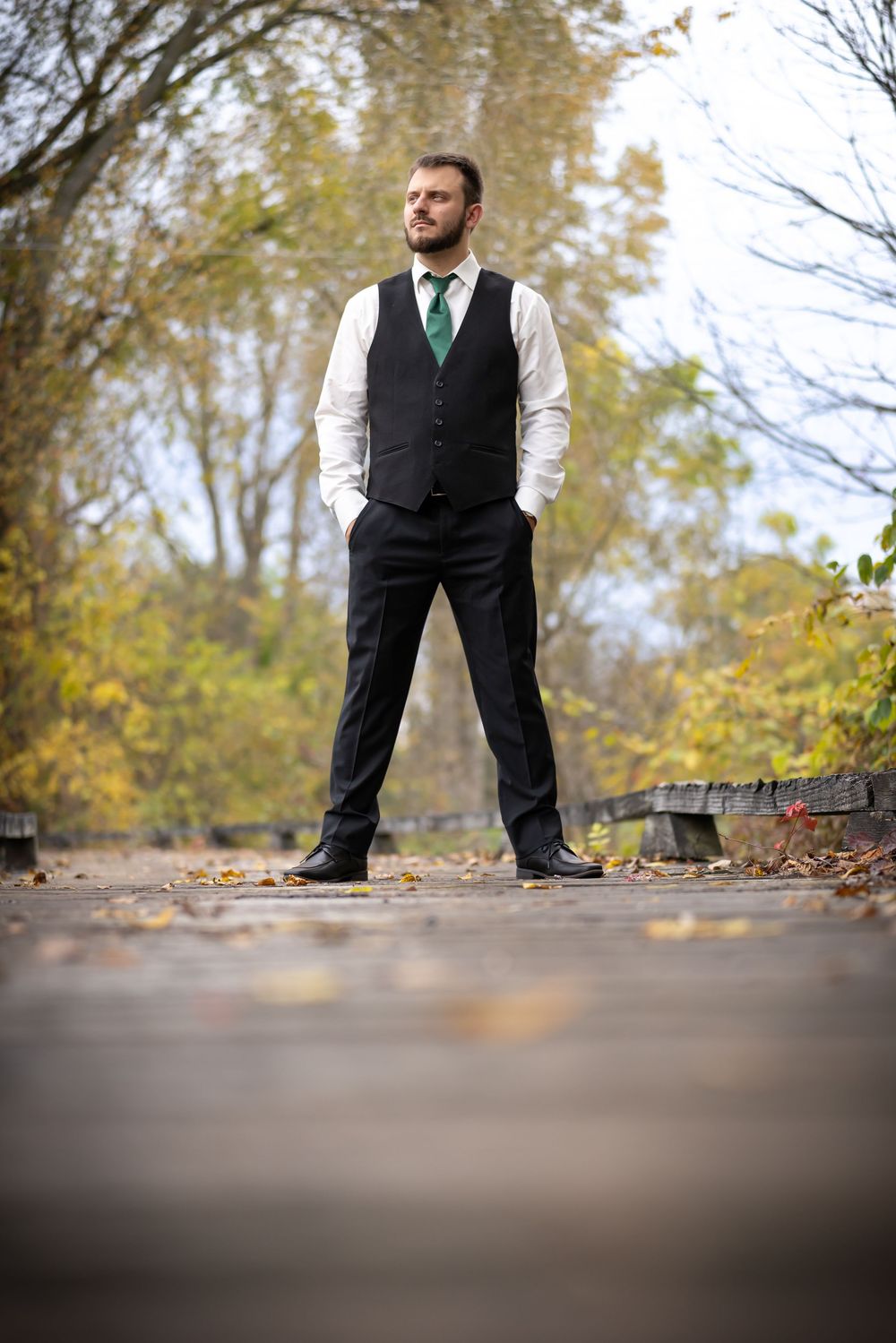 A groom in formal attire with a green tie poses outdoors among autumn trees.