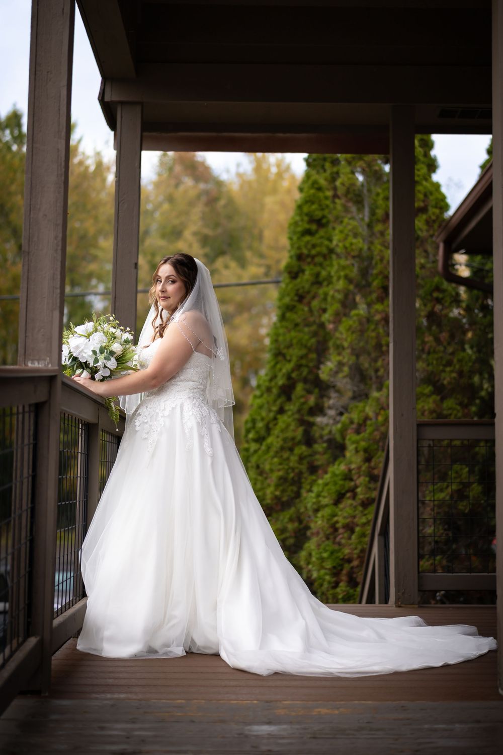 A bride in a flowing white wedding gown poses gracefully on a covered wooden deck surrounded by greenery.