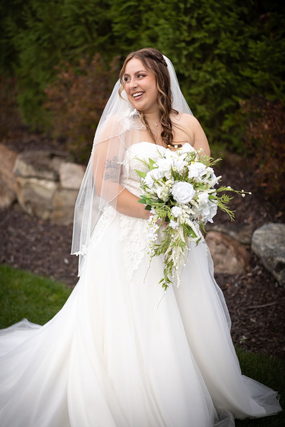 A bride in a flowing white gown holds a stunning white and green floral bouquet against a natural backdrop.