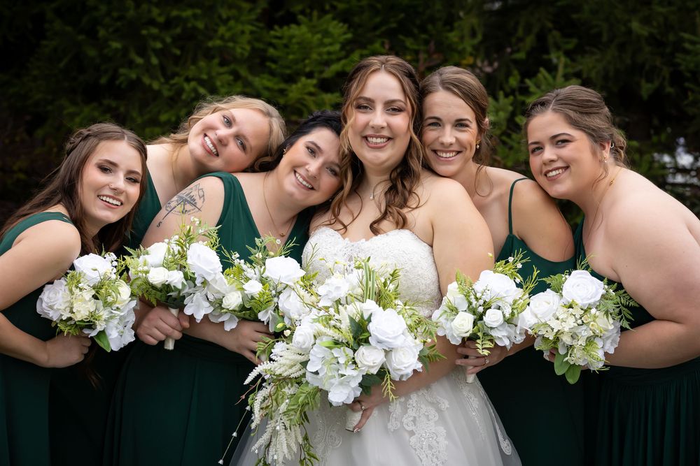 A group in emerald green dresses and white floral bouquets pose together outdoors for a wedding photo.