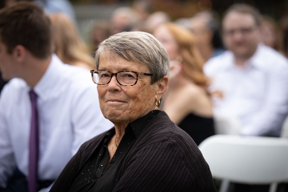 A guest in black attire and glasses watches attentively during an outdoor ceremony.