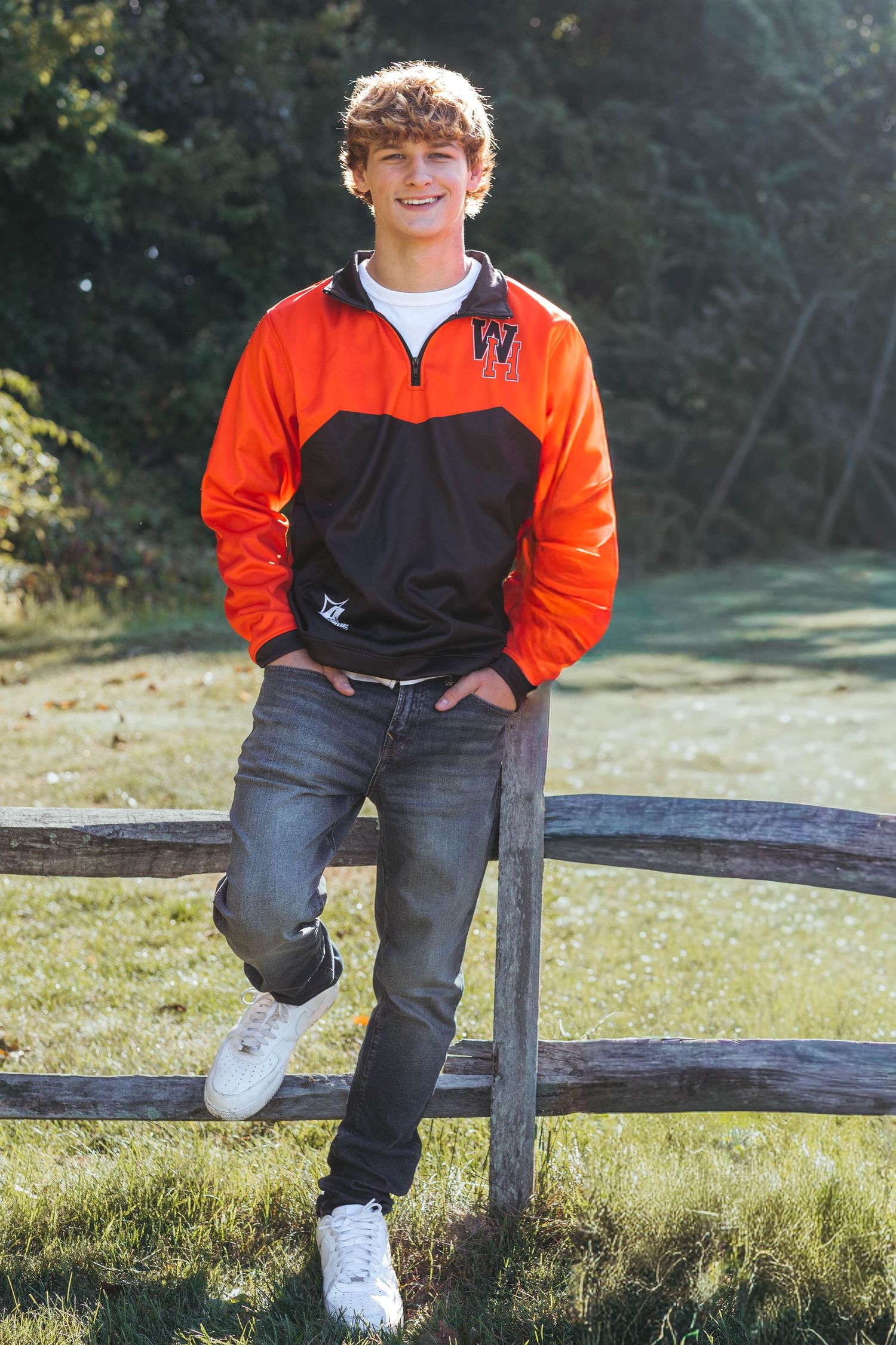 Person in orange and black jacket sitting casually on wooden fence in outdoor setting.