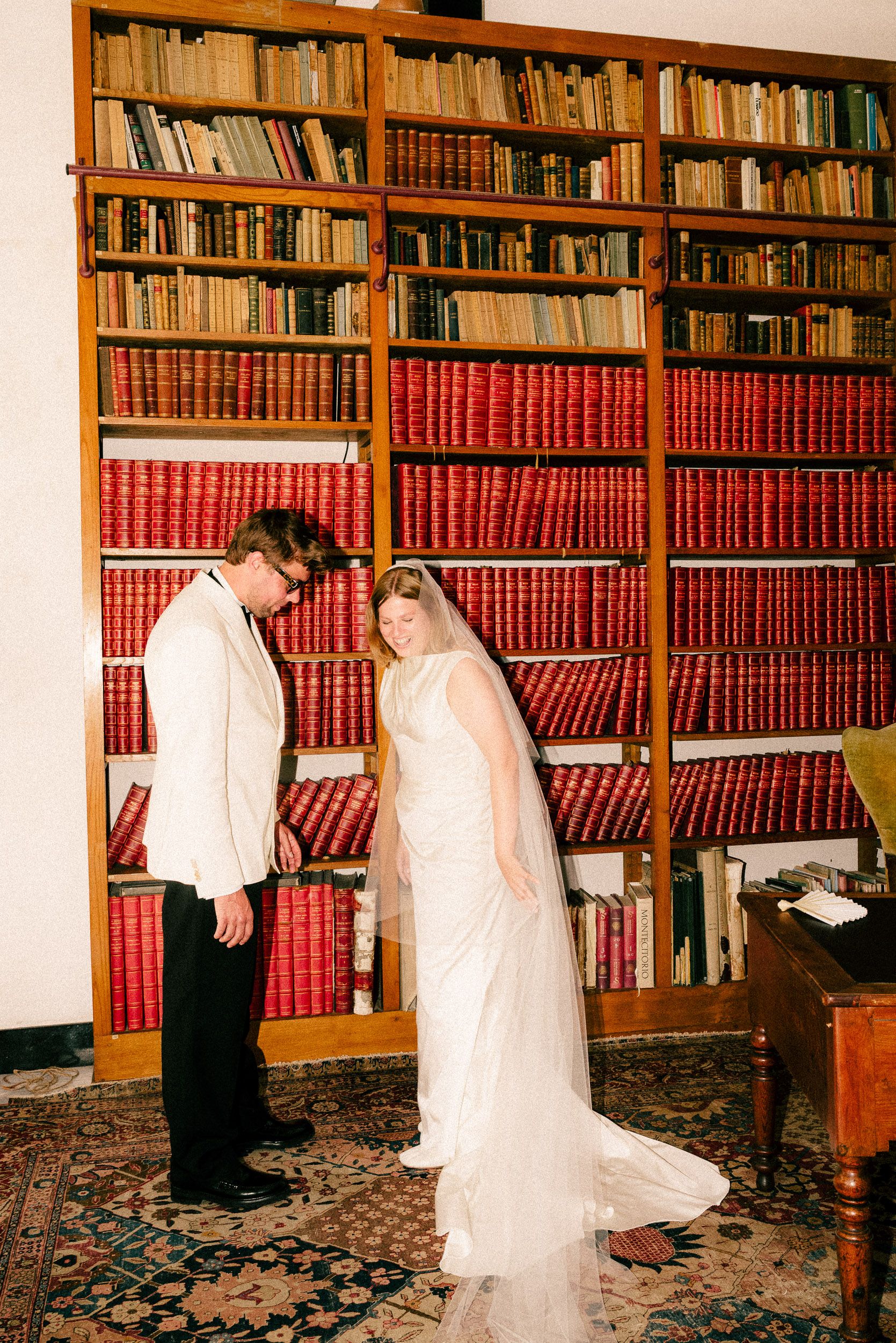 A wedding couple browses through books in a library with wooden shelves and red-bound volumes.