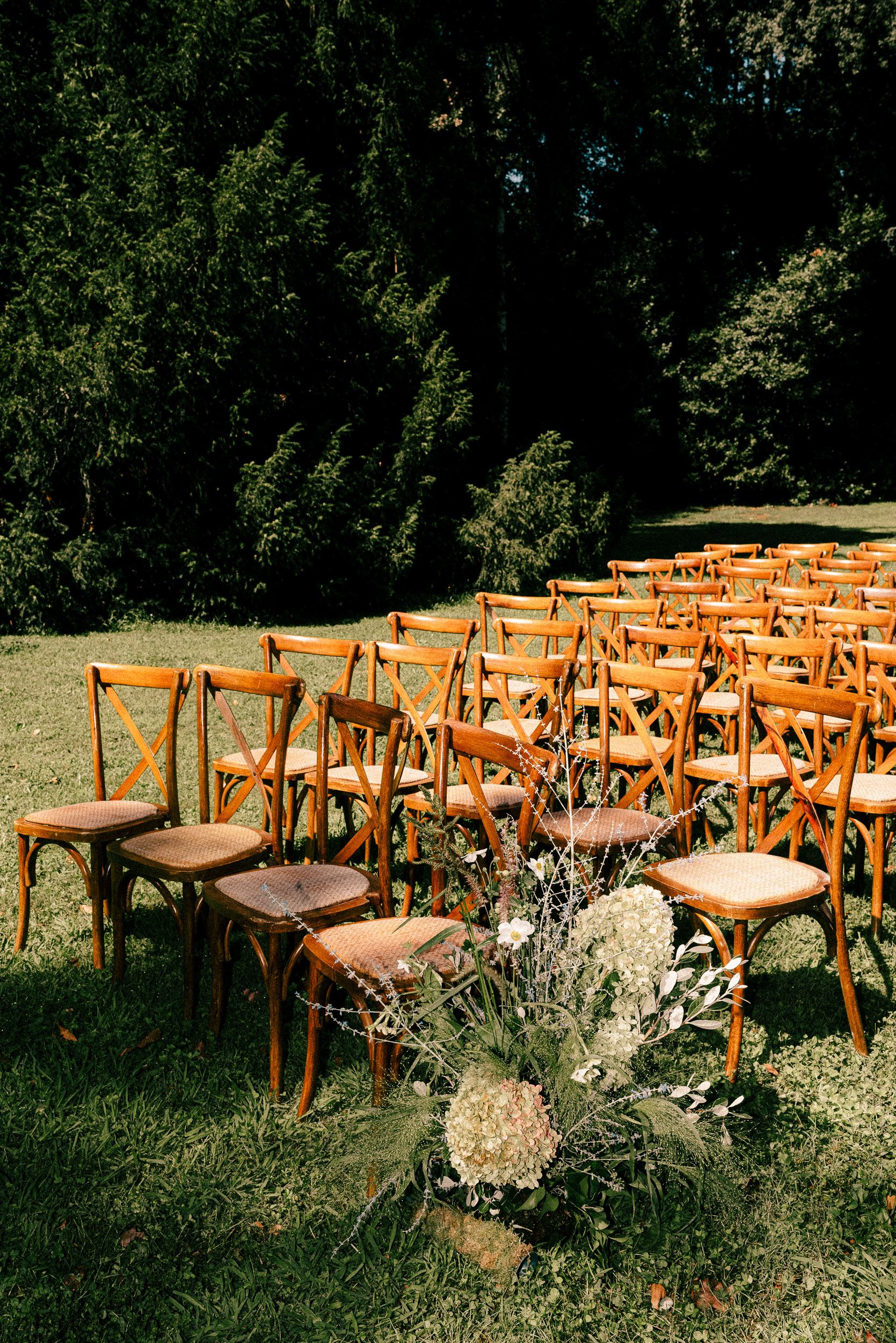 Rustic wooden crossback chairs arranged for an outdoor wedding ceremony with a natural dried floral arrangement on the aisle.