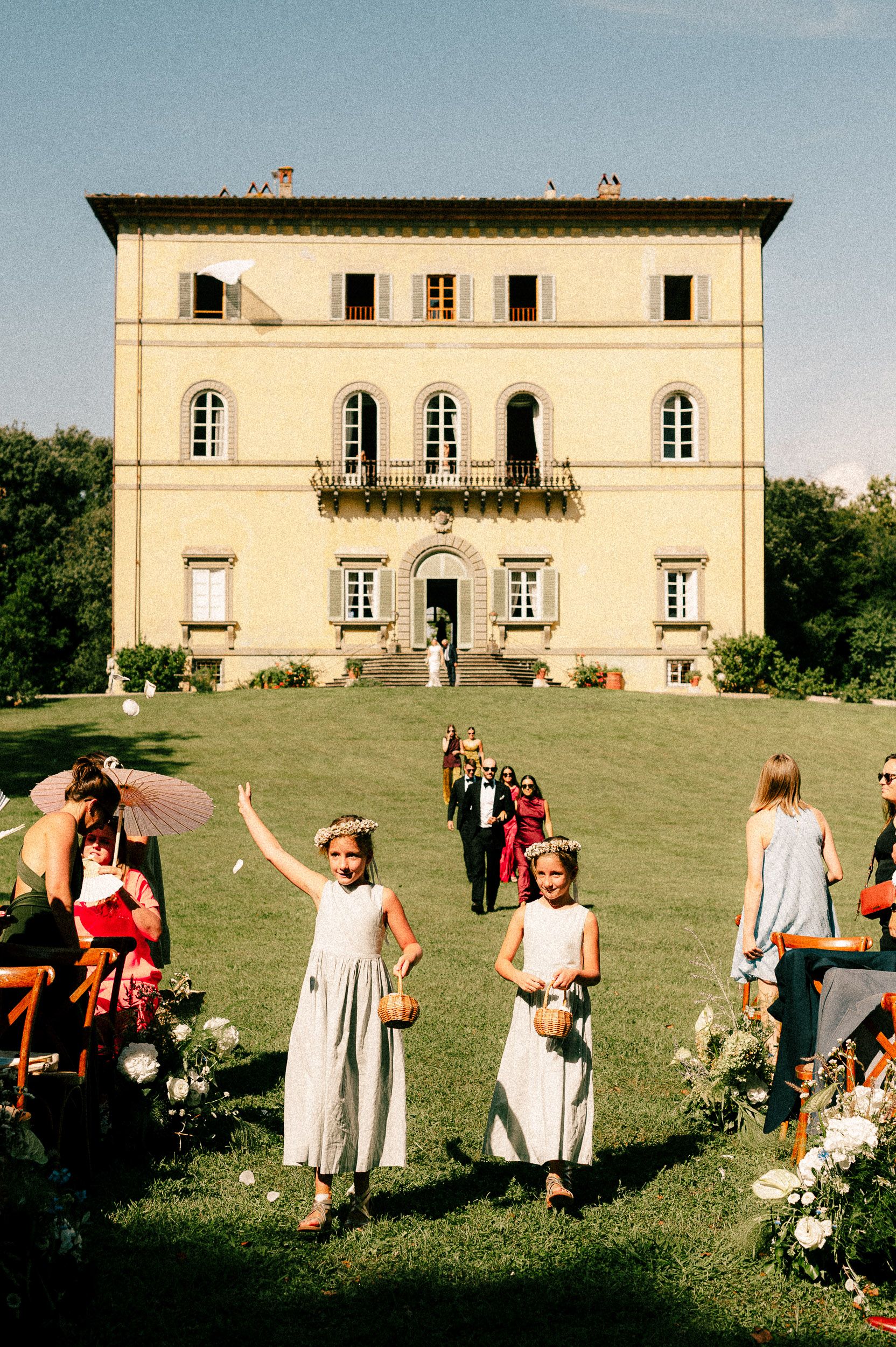 People gather on a sunny lawn in front of an elegant Italian villa for what appears to be a festive outdoor celebration.