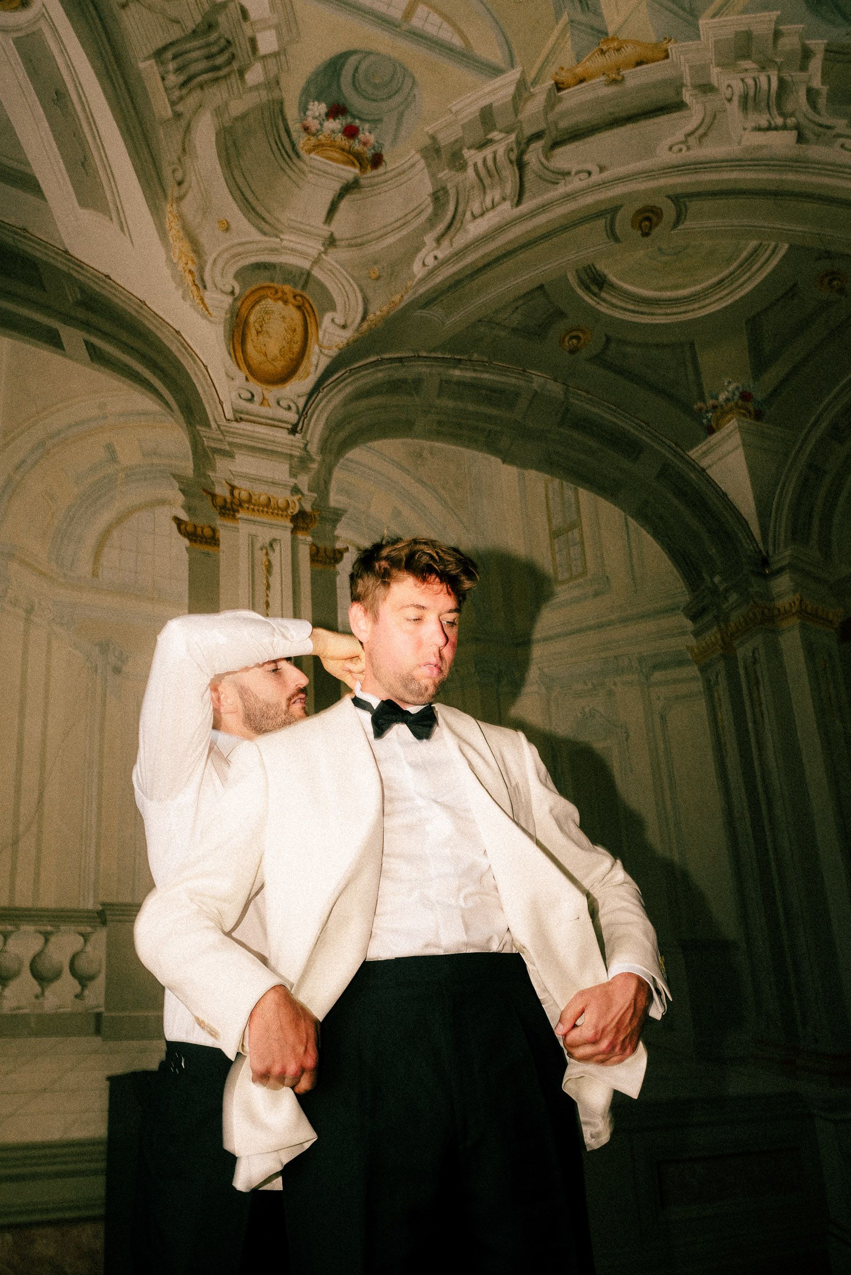 A person in formal white tuxedo jacket and black bow tie poses dramatically in an ornate baroque-style room.