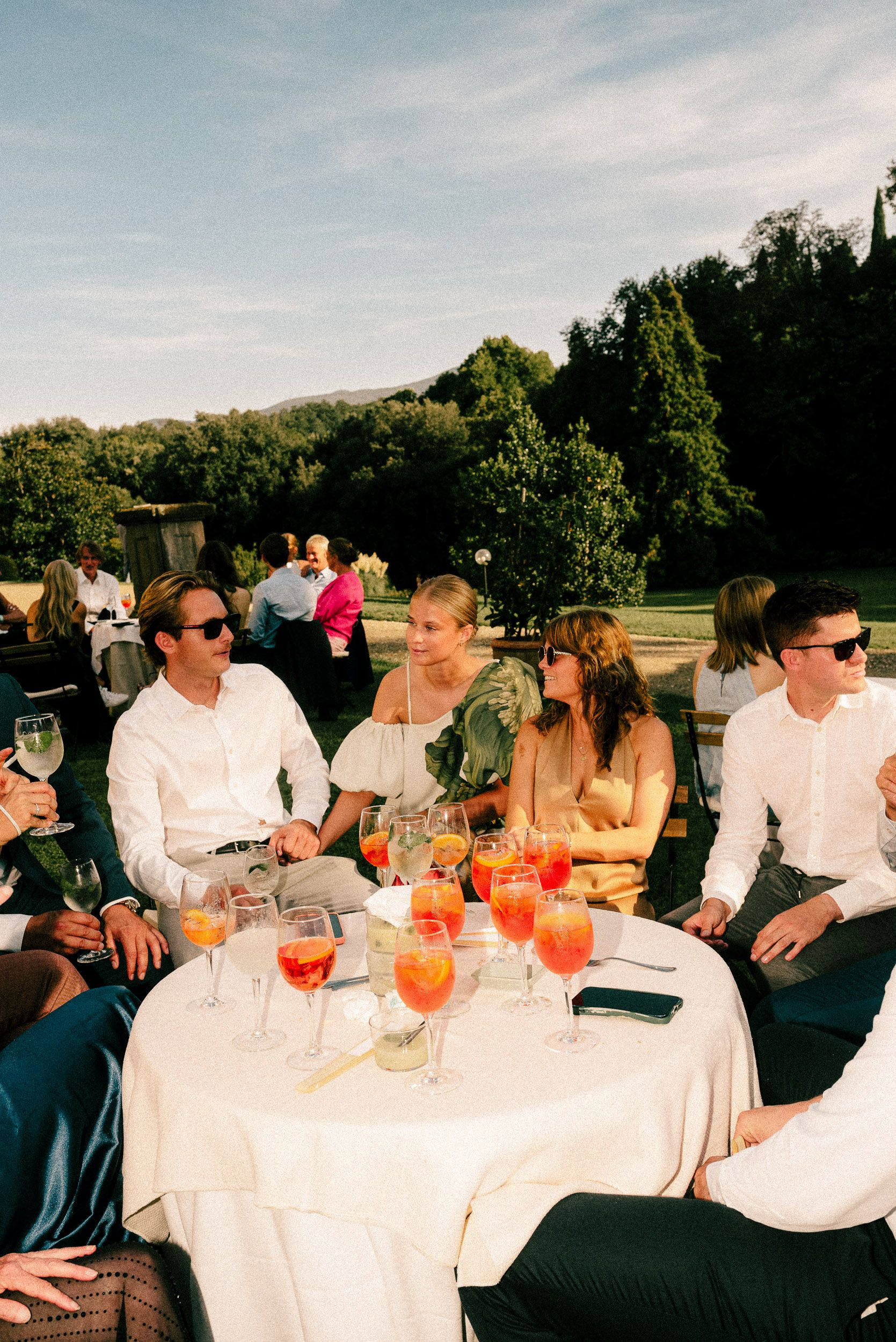 Group of people enjoying drinks at an outdoor party with white tablecloths and orange cocktails at sunset.