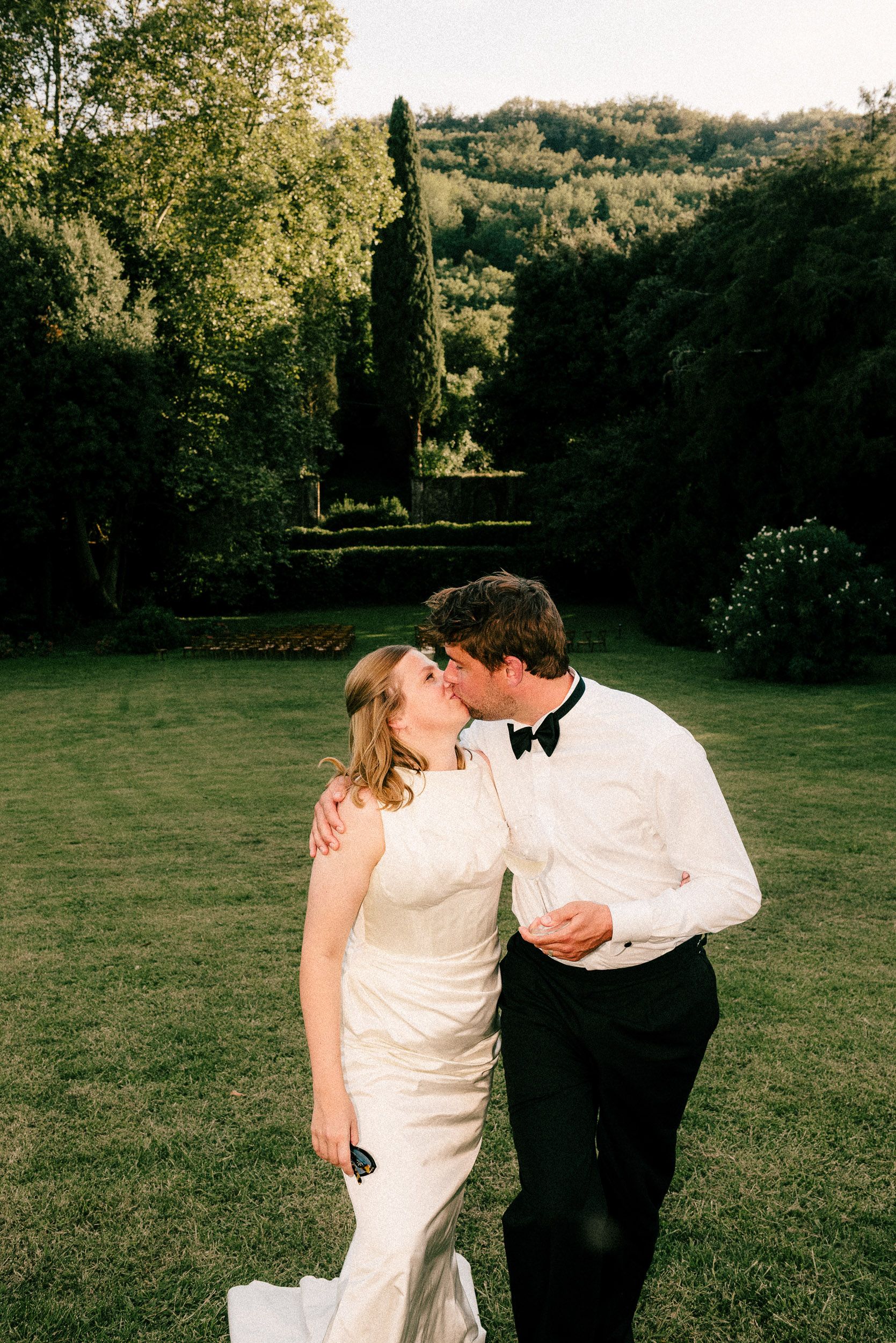 A couple shares a romantic moment in formal wedding attire on a manicured garden lawn with cypress trees in background.