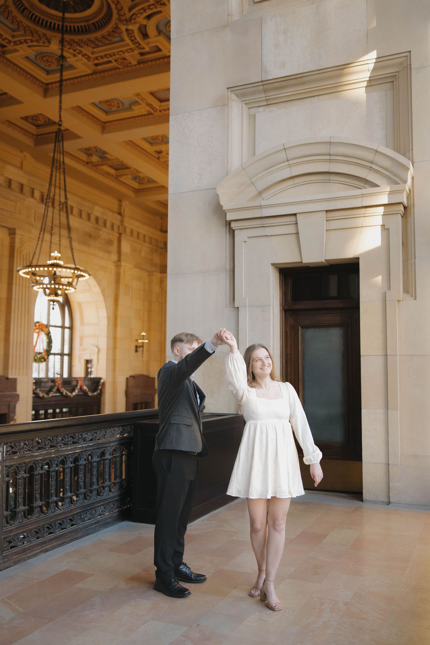 Two figures in formal attire interact in an ornate hallway with decorative ceilings and architectural details.