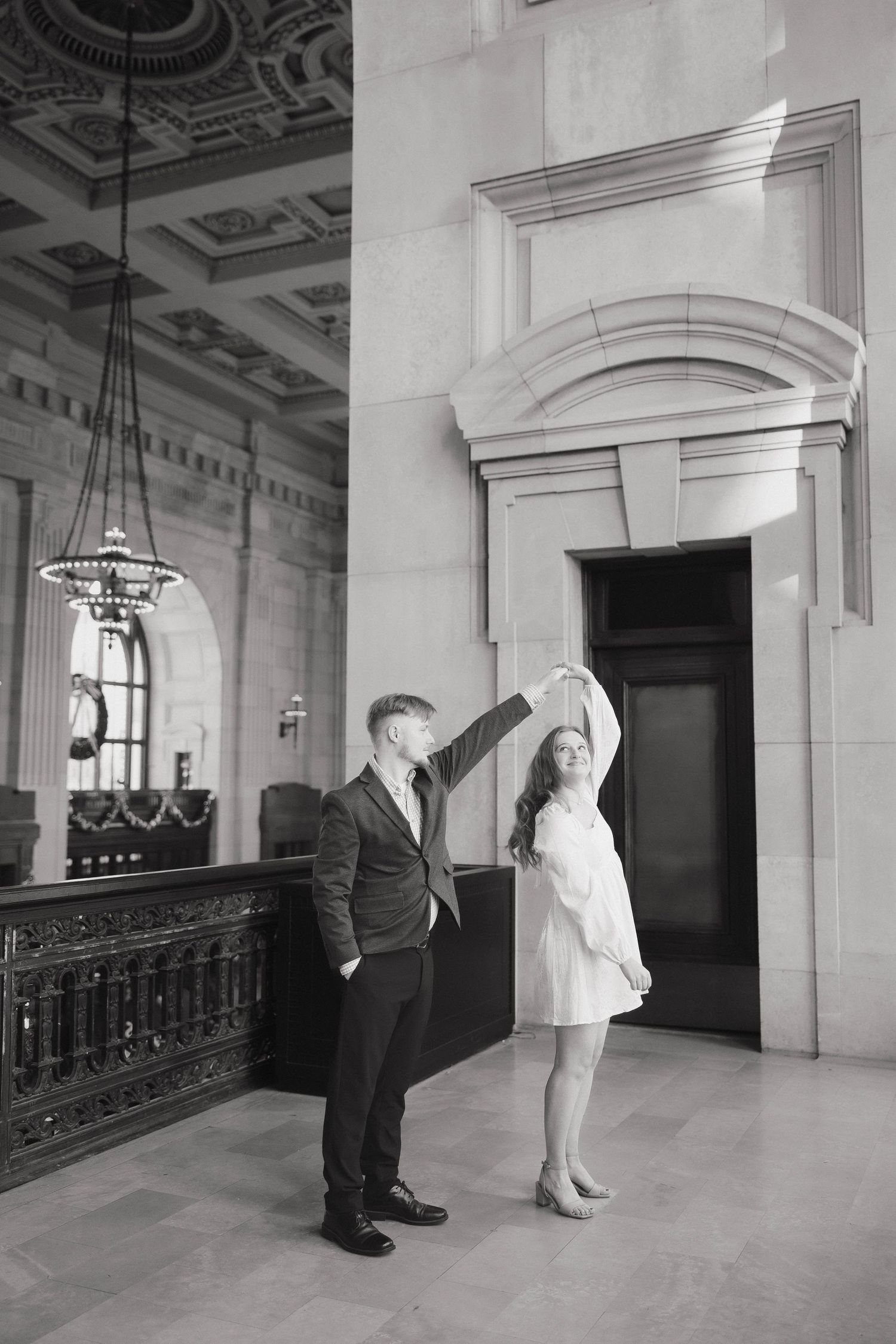 Two people in formal attire stand in an ornate hallway with decorative architectural details and lighting fixtures.