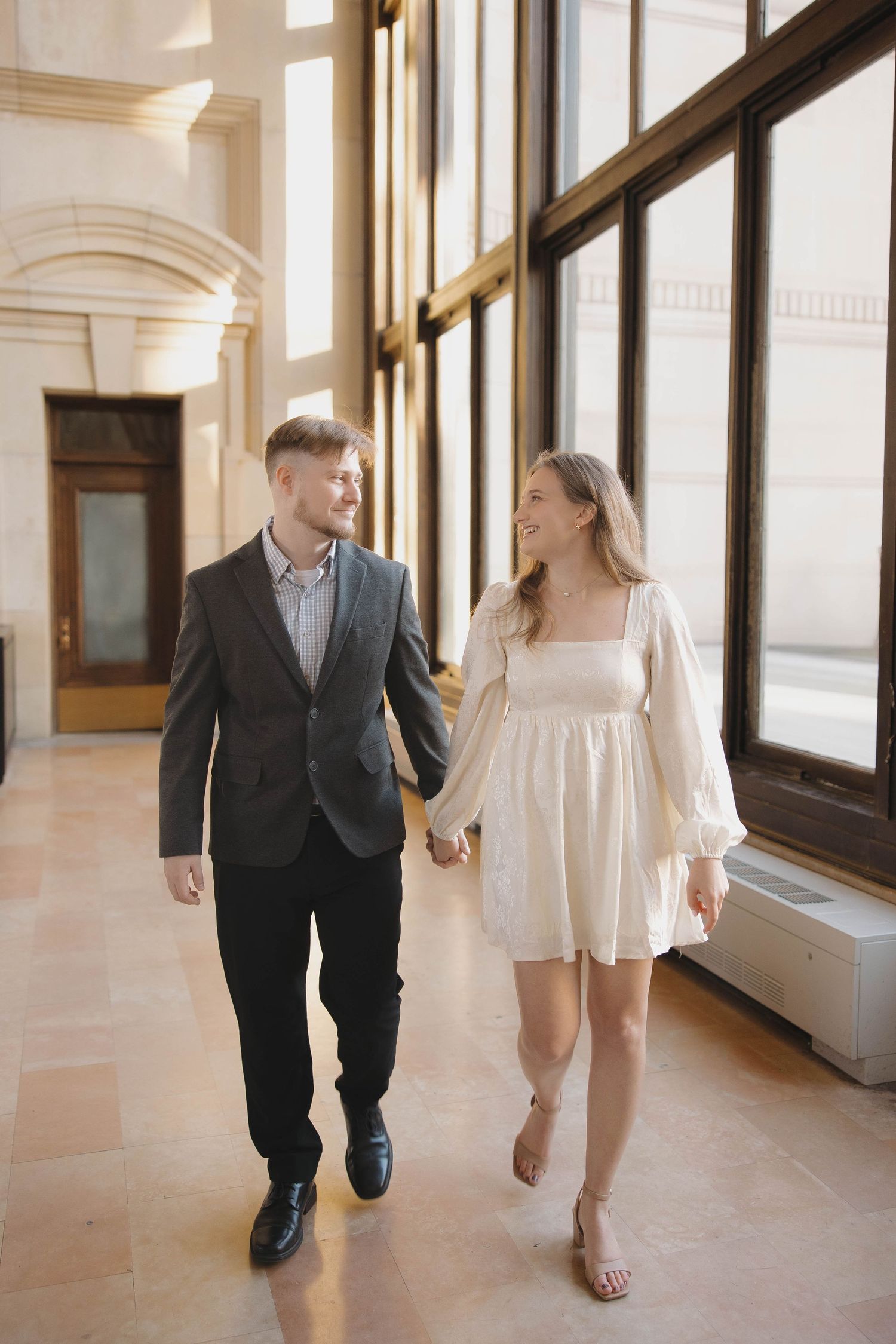 A couple walks hand in hand through an elegant hallway with arched windows and marble floors.