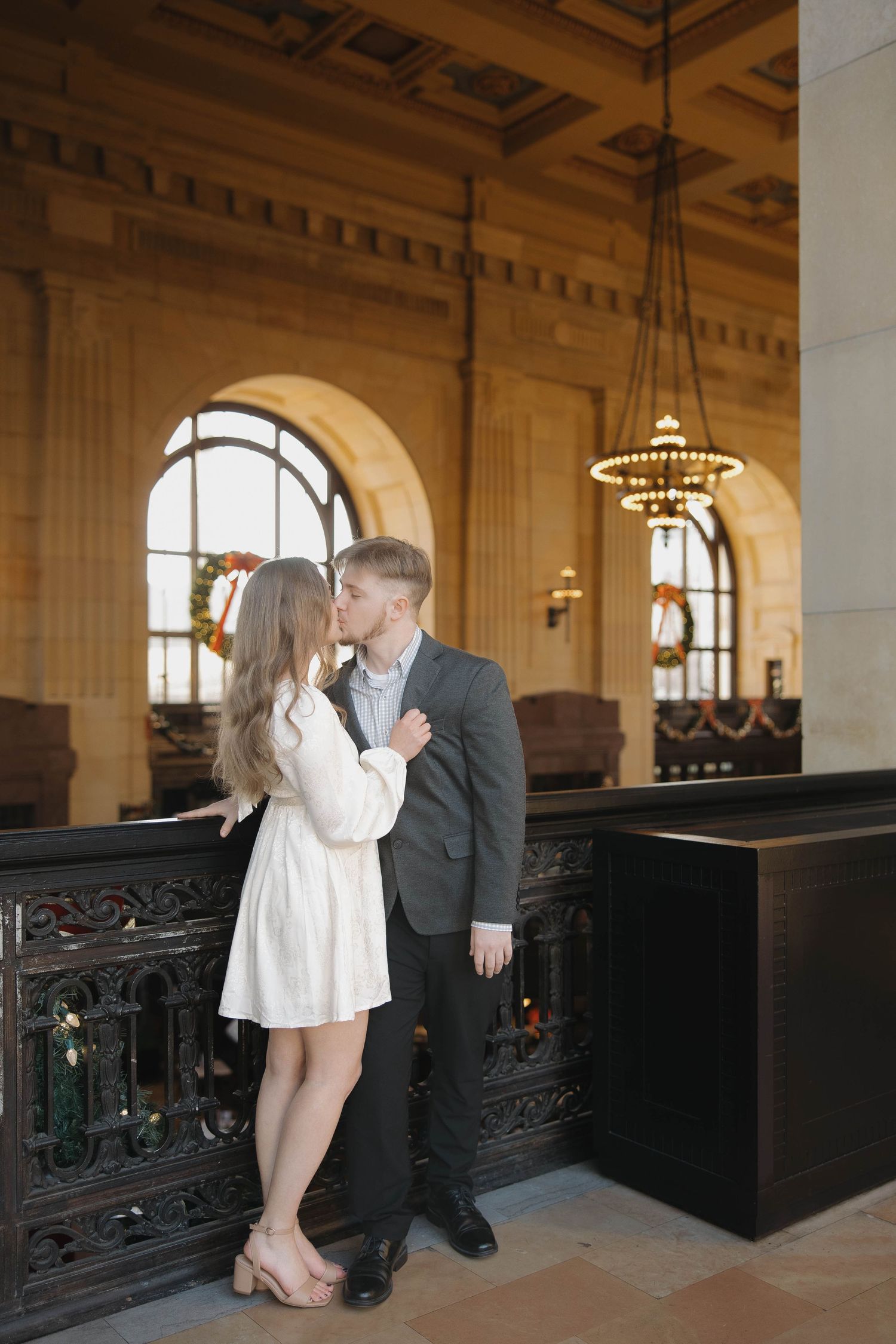 A romantic couple shares a tender moment in an elegant historic building with arched windows and ornate architectural details.