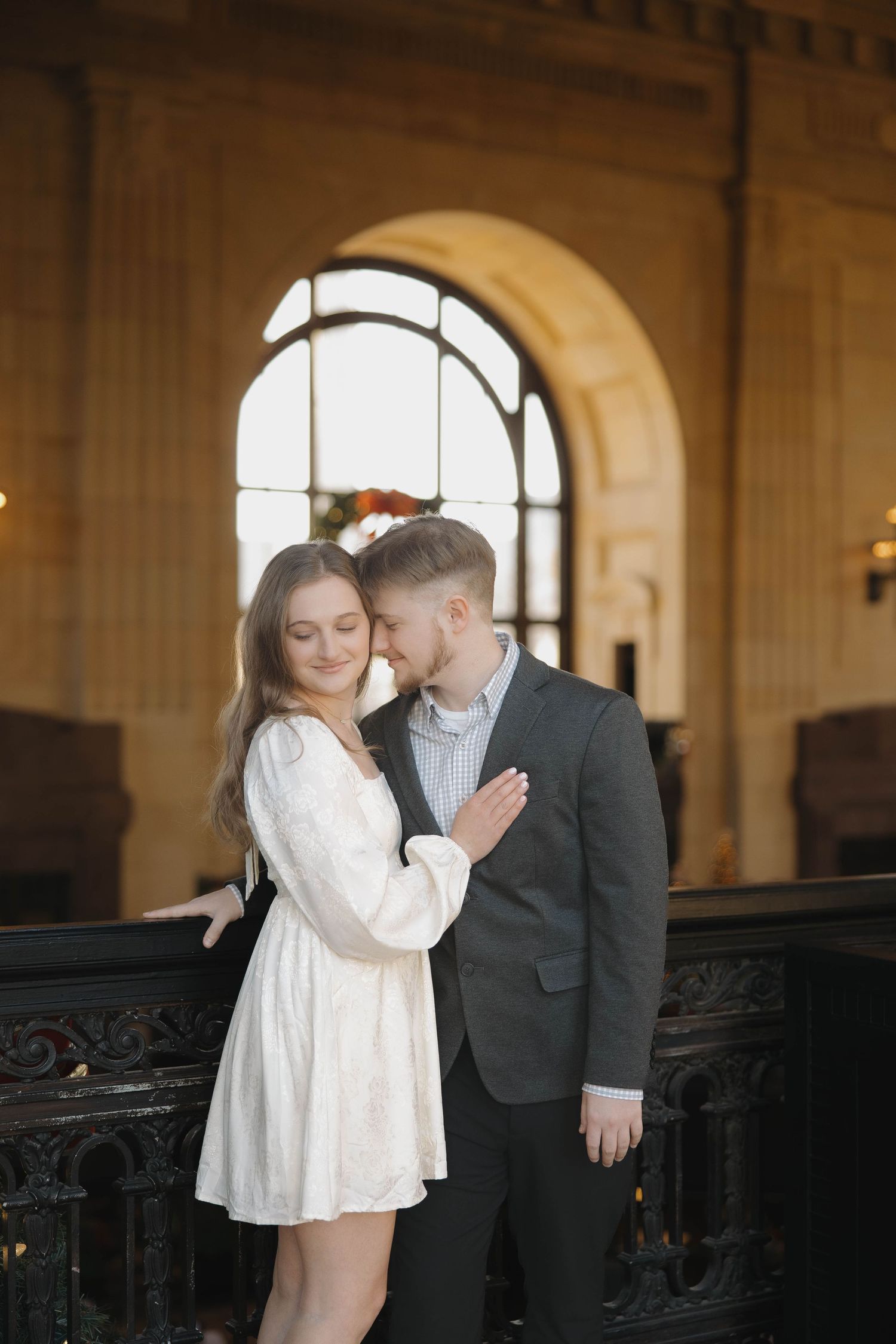 A romantic engagement photo session in an elegant indoor venue with ornate black railings and arched windows.