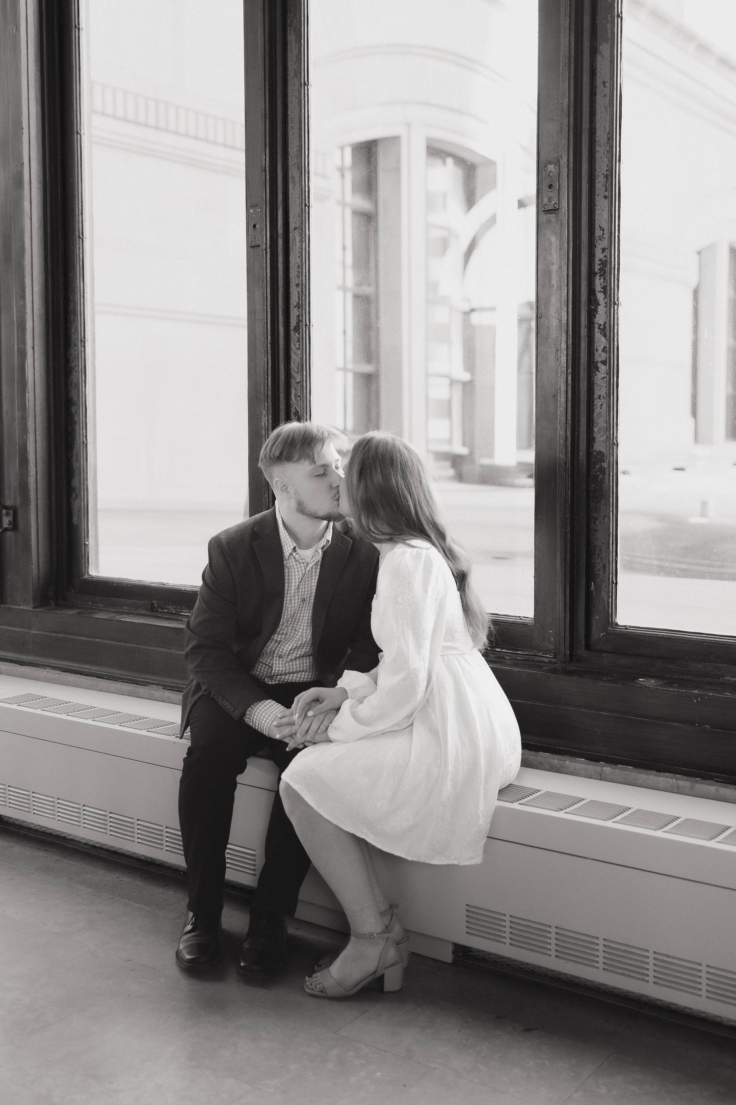 A couple shares an intimate moment by large windows in a historic building with natural light streaming in.