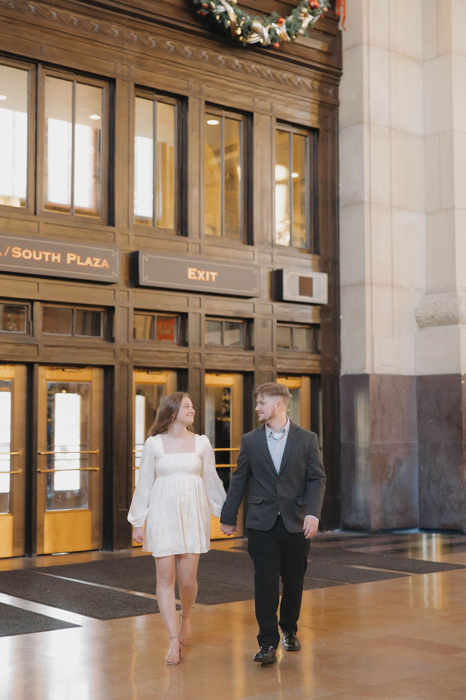 A couple walking through a grand historic building hallway with EXIT signs and wooden details.