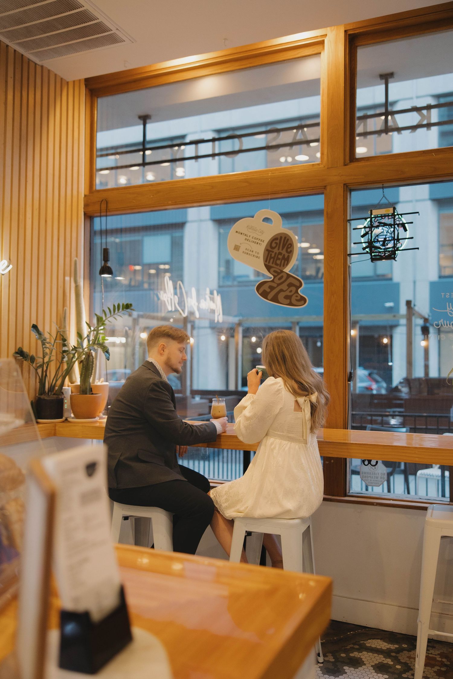 Two people sit at a modern wooden bar counter with large windows and ambient lighting in a contemporary setting.