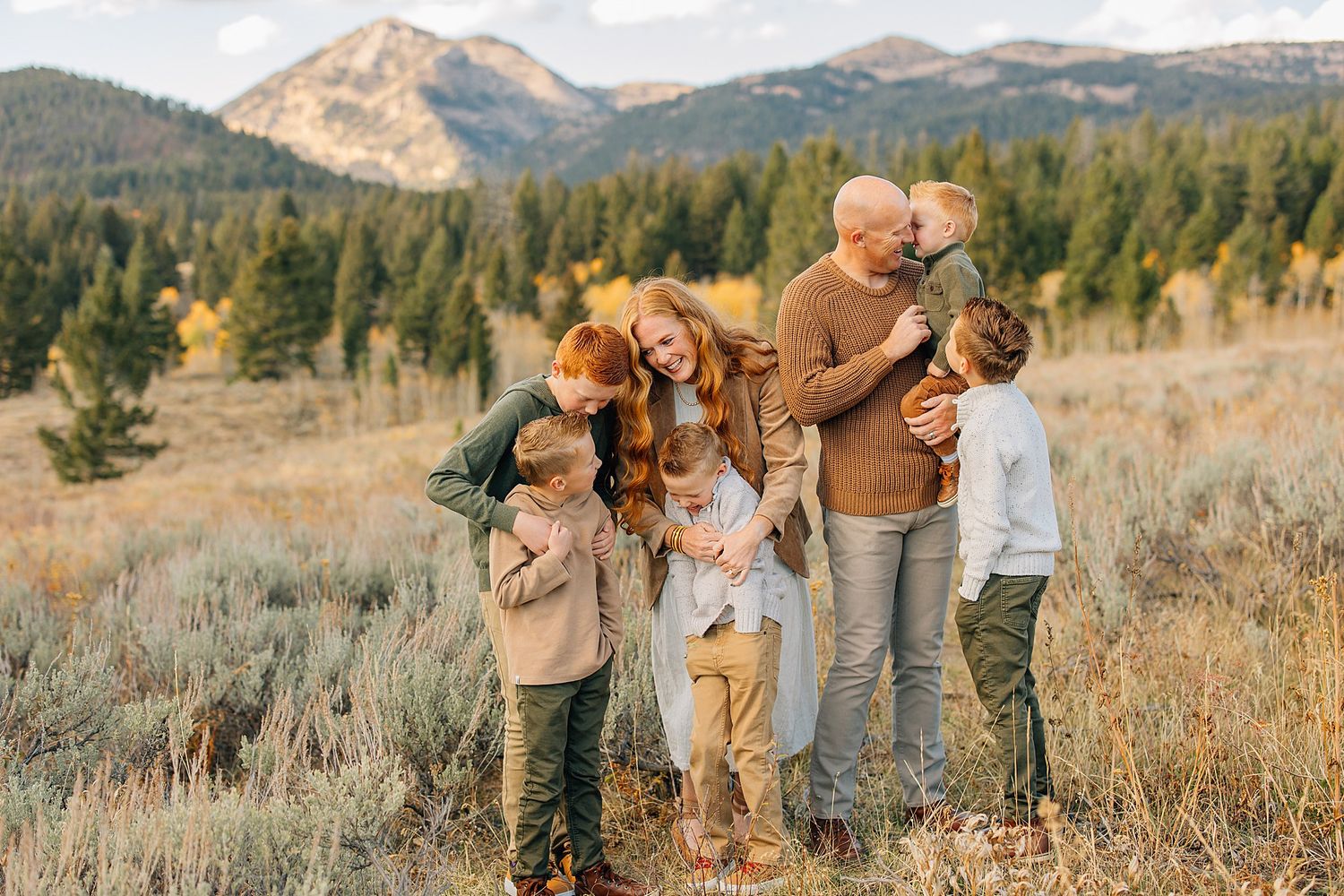 A family in earth-toned outfits stands together in a golden mountain meadow during autumn.