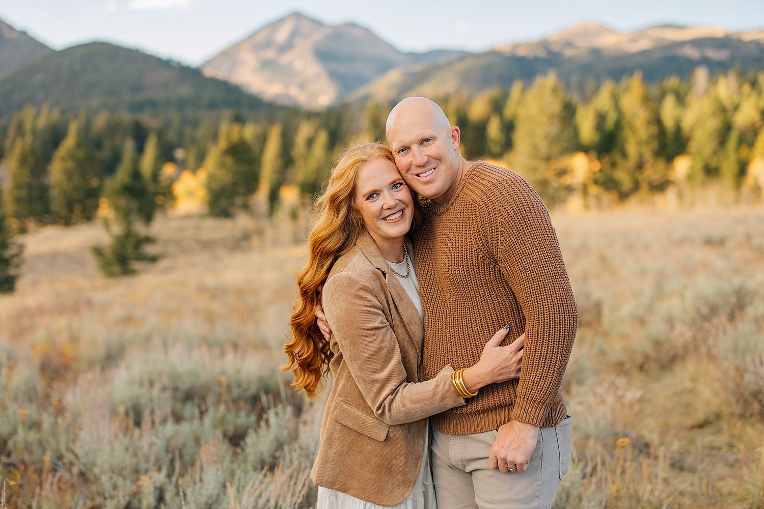 Family wearing brown and beige outfits poses together in scenic mountain landscape during autumn.