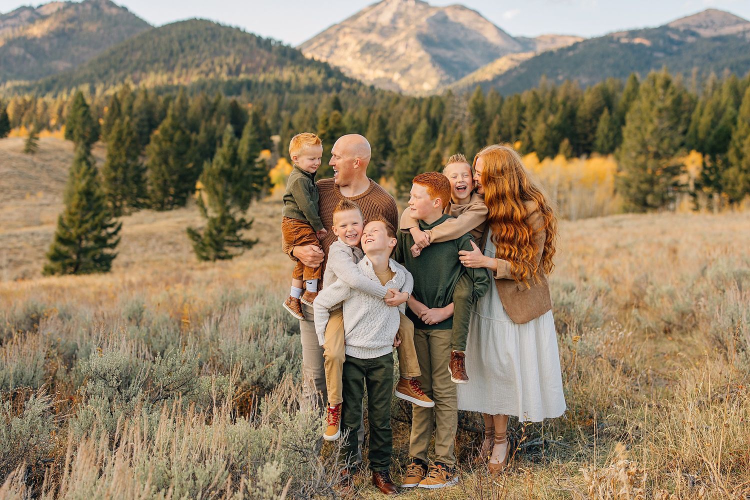 A group gathers in a golden mountain meadow during sunset with evergreen trees and peaks in the background.