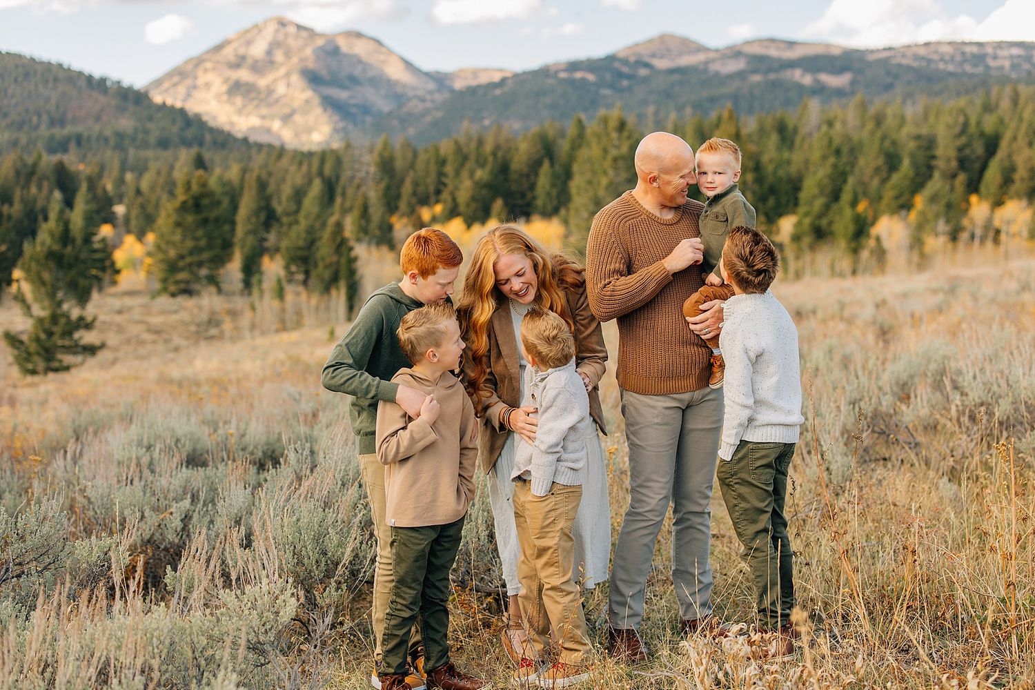 A family group poses together in a golden autumn field with snow-capped mountains and evergreen forest in the background.
