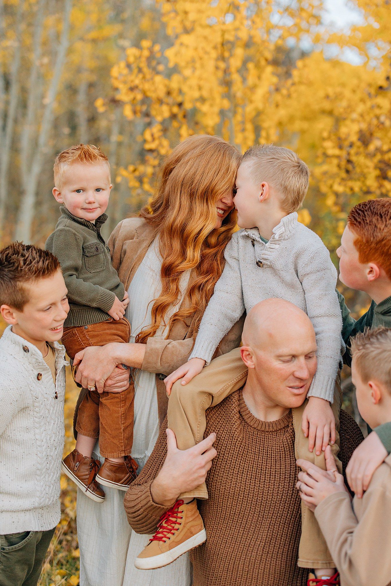 A family wearing earth-toned clothing embraces and plays together against a backdrop of golden autumn trees.