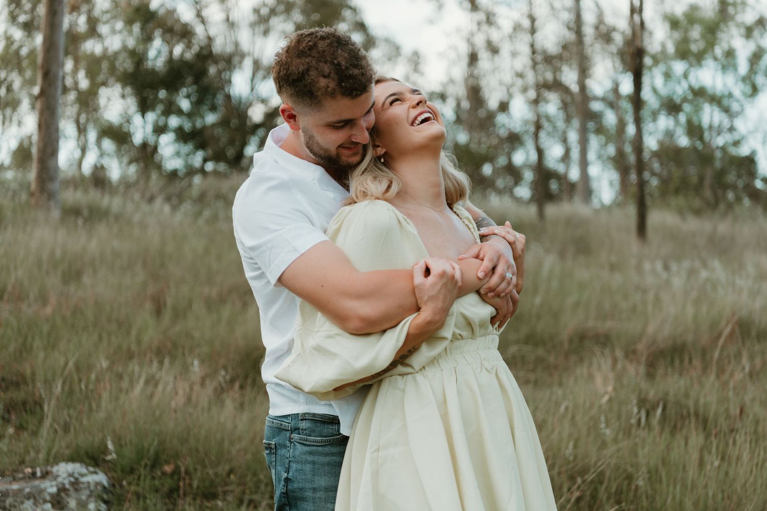 Happy couple embracing outdoors in a grassy field, woman in yellow dress laughing joyfully.