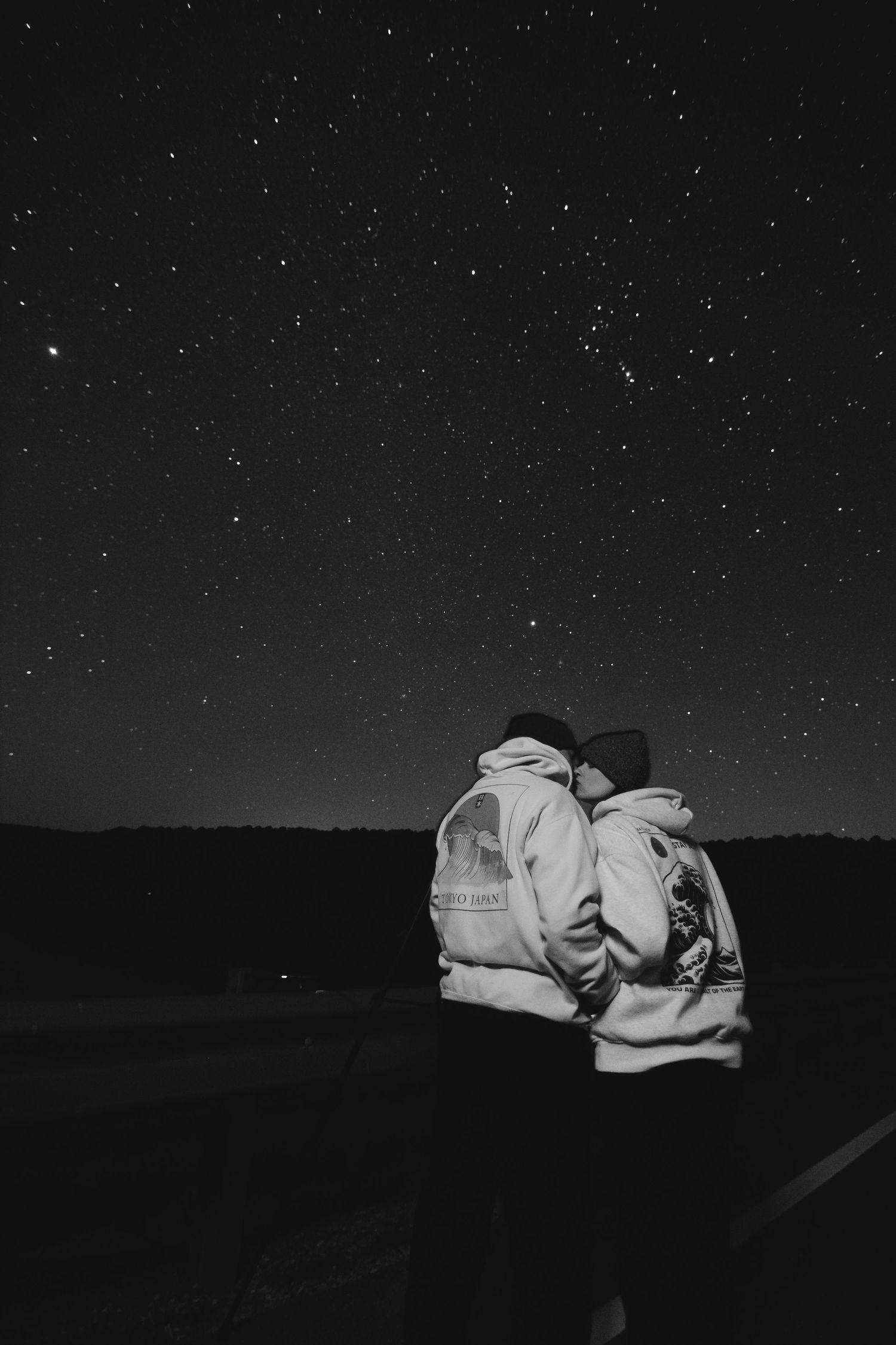 A starry night sky stretches over dark a romantic couple with rolling hills in a black and white panoramic view.