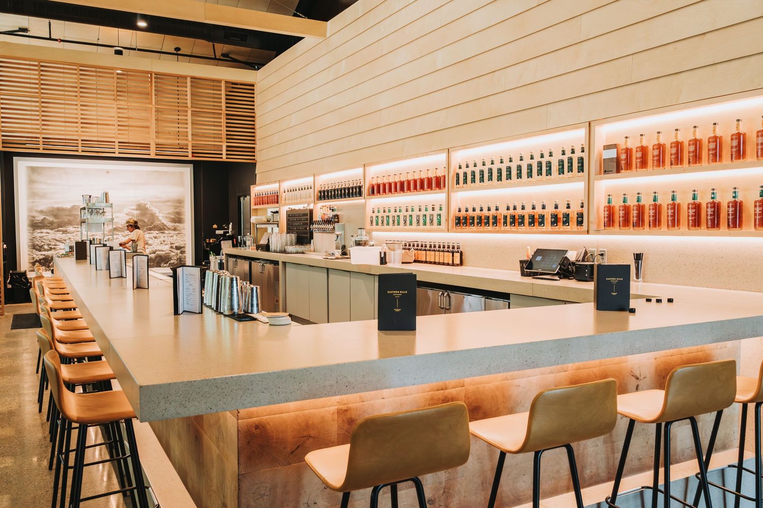 Modern bar interior with sleek counter seating and rows of illuminated bottles displayed on wooden wall shelving.