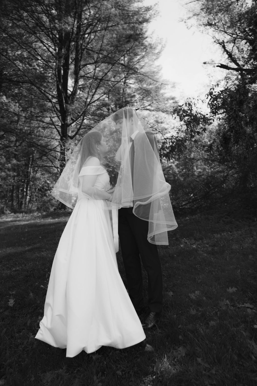 A couple in wedding attire stands together in a wooded outdoor setting captured in black and white photography.