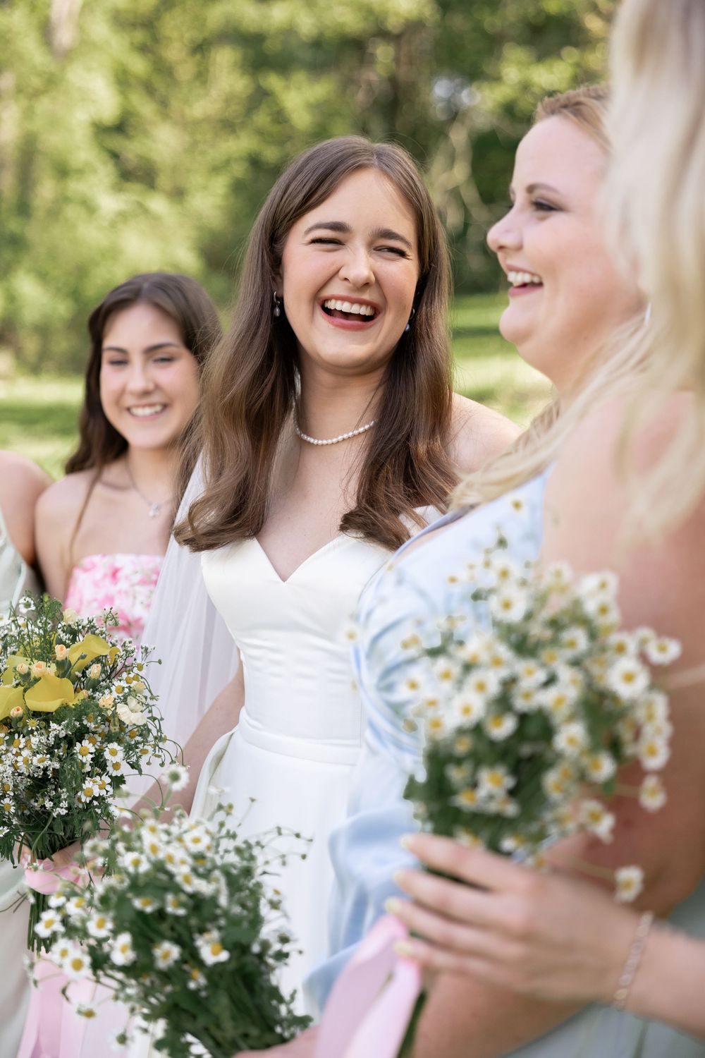 Bridesmaids in white dresses share joyful laughter while holding delicate wildflower bouquets outdoors.
