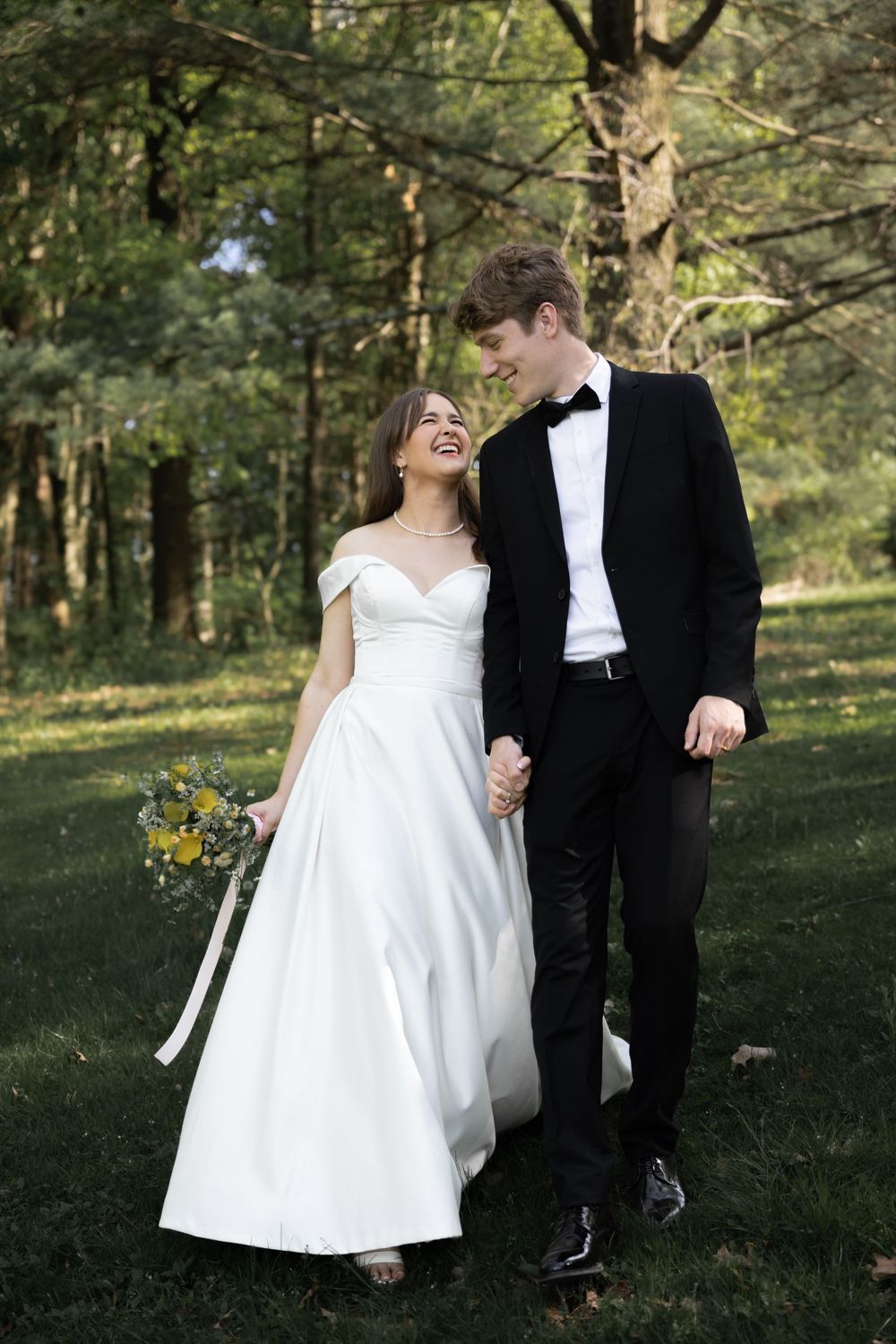 A joyful couple in wedding attire share a candid moment while walking together in a sunlit wooded area.