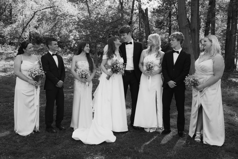 A formal wedding party in black and white poses together outdoors in a garden setting.