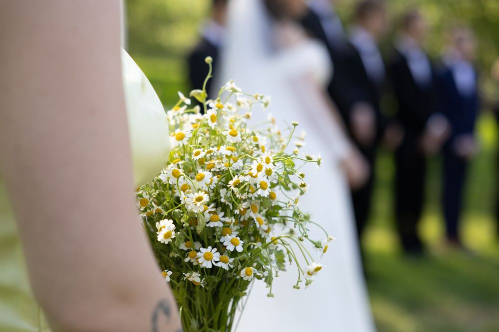 A delicate bouquet of white flowers with small daisies and baby's breath being held during an outdoor wedding ceremony.