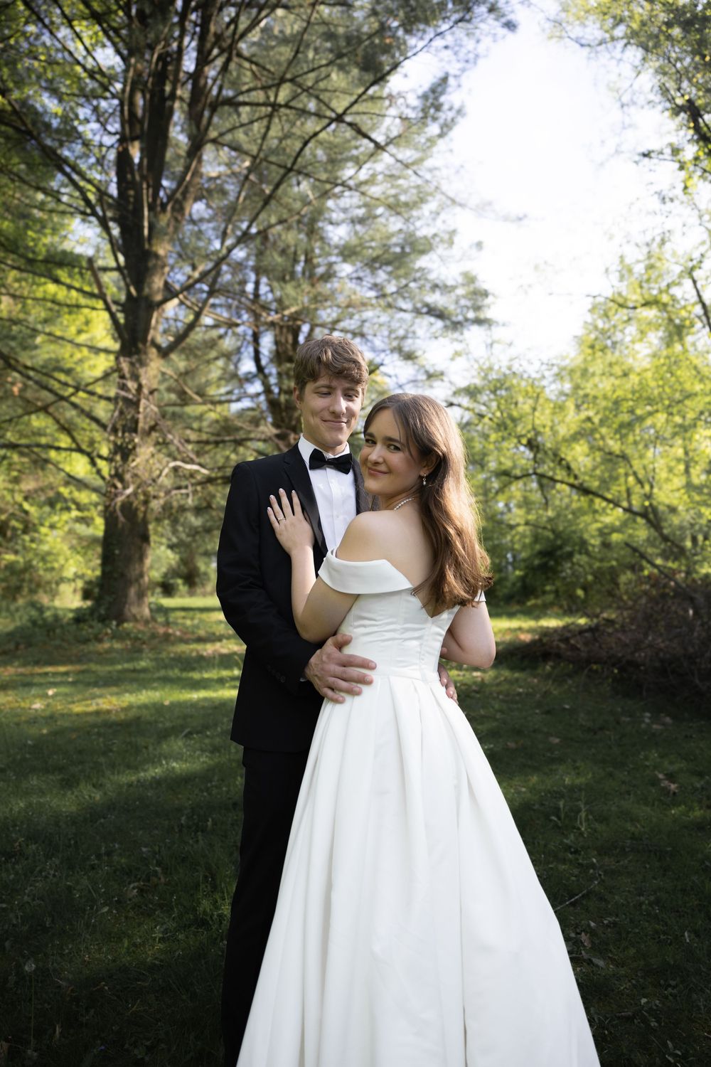 A couple in formal wedding attire embraces while standing in a lush green park setting during their special day.