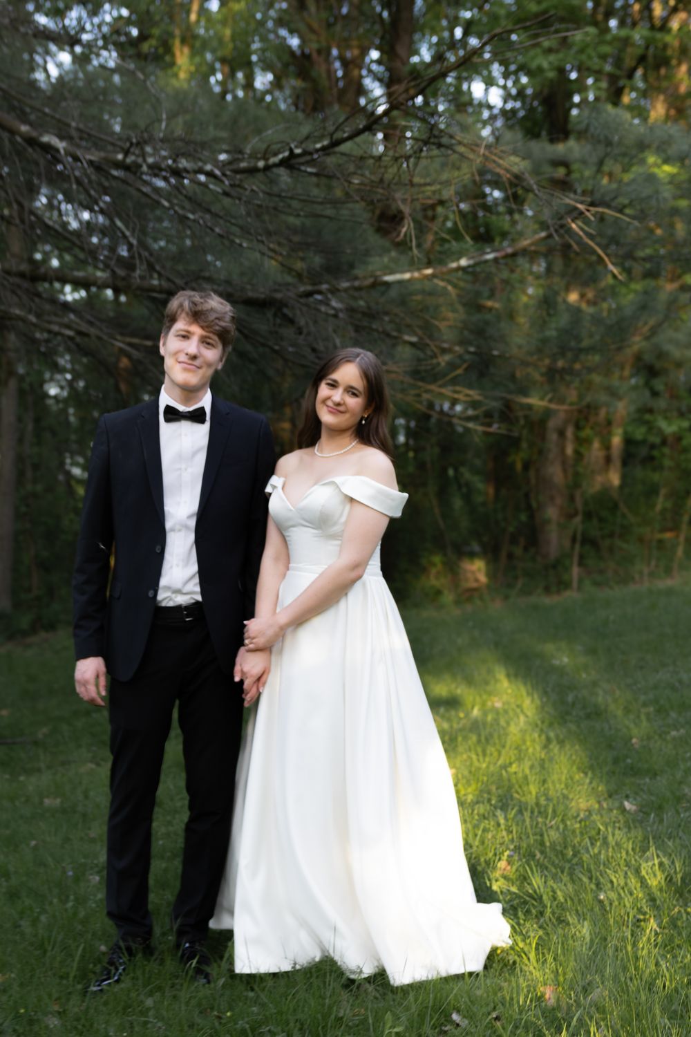 A bride and groom stand together in a wooded outdoor setting during their wedding photo session at sunset.