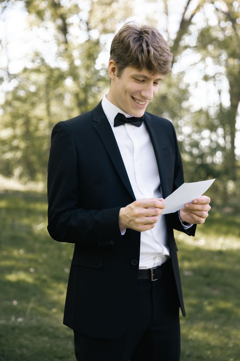 A person in a black tuxedo and bowtie reads a letter while standing outdoors in natural lighting.