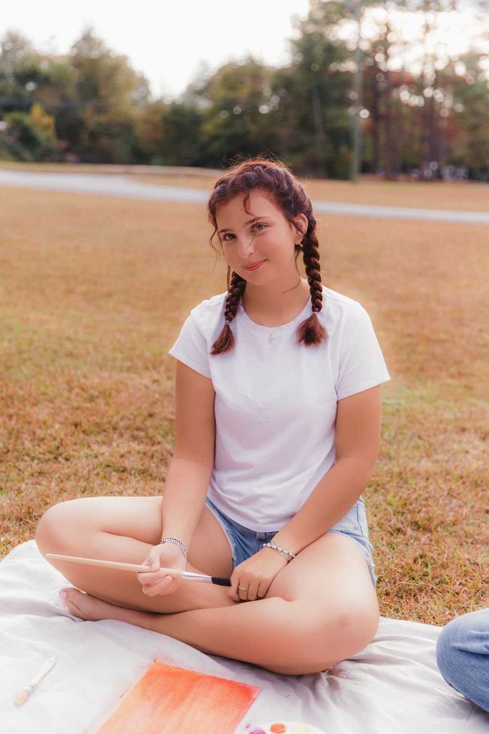 Student in white t-shirt and denim shorts sits cross-legged on blanket outdoors during autumn afternoon.