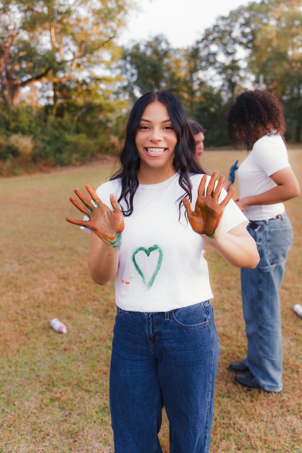 A person in a white t-shirt and overalls shows their hands covered in green paint while standing in a grassy field.