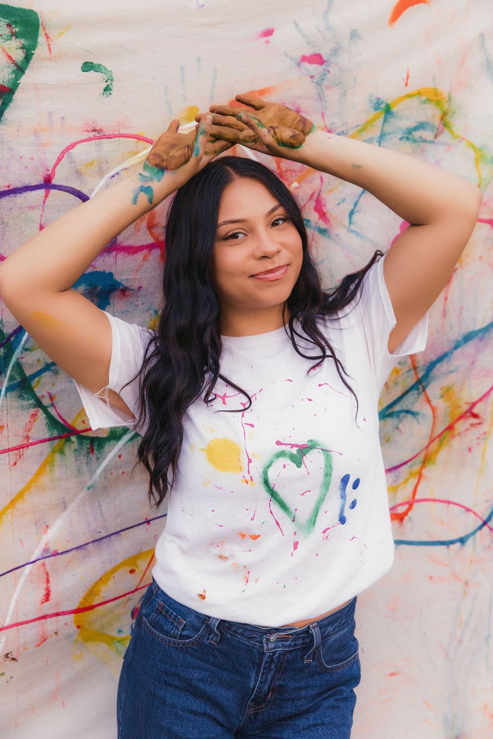 Person in white shirt and jeans poses playfully against wall with vibrant paint splatter art.