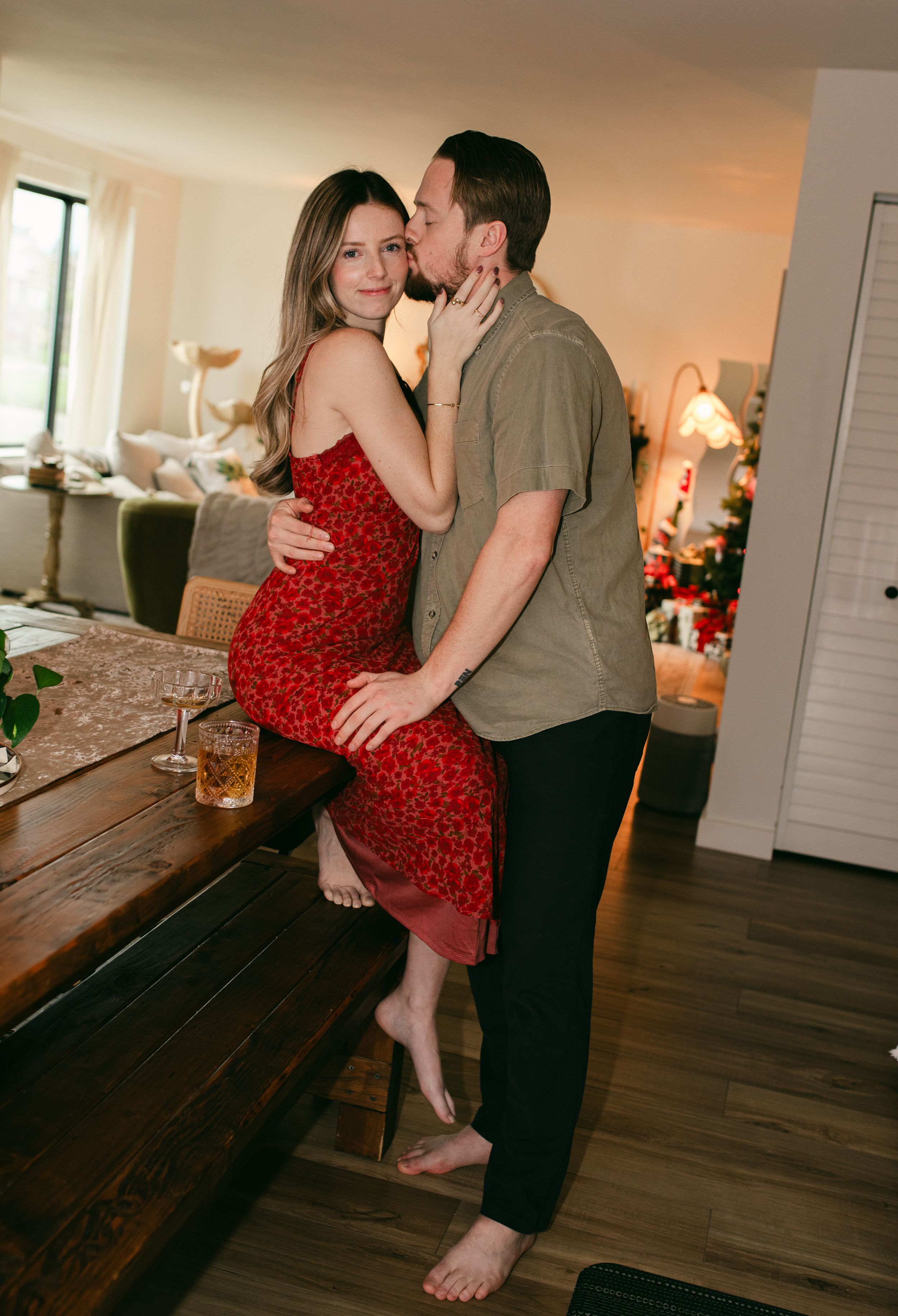 A romantic couple embraces in their living room, with her wearing a red dress and him in casual attire.