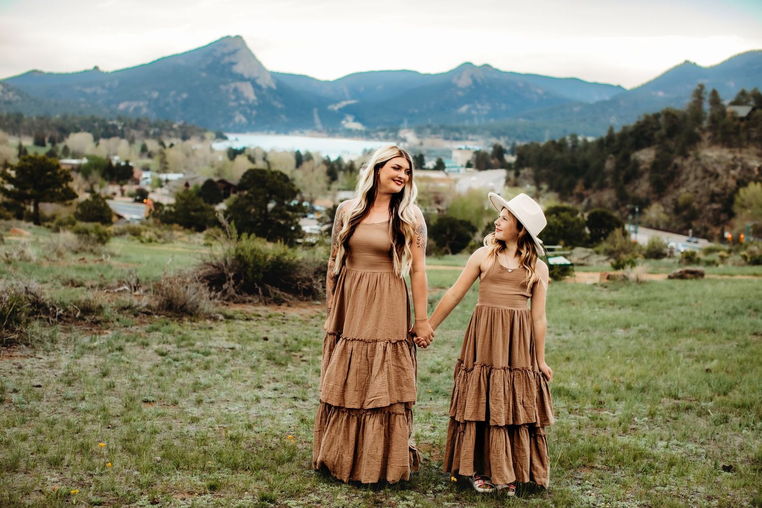 Two people in matching brown tiered maxi dresses stand holding hands in a mountain meadow landscape.