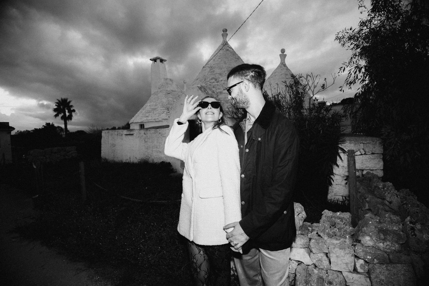 A couple stands together in front of traditional trulli houses in Alberobello, Italy on a cloudy evening.