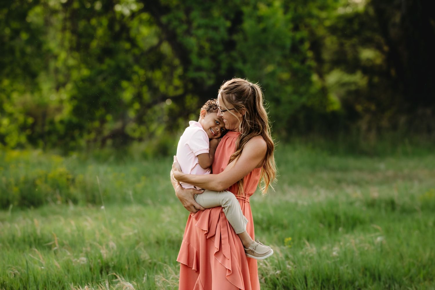 A tender embrace shared between two figures in a coral dress and white outfit standing in a sunlit meadow.