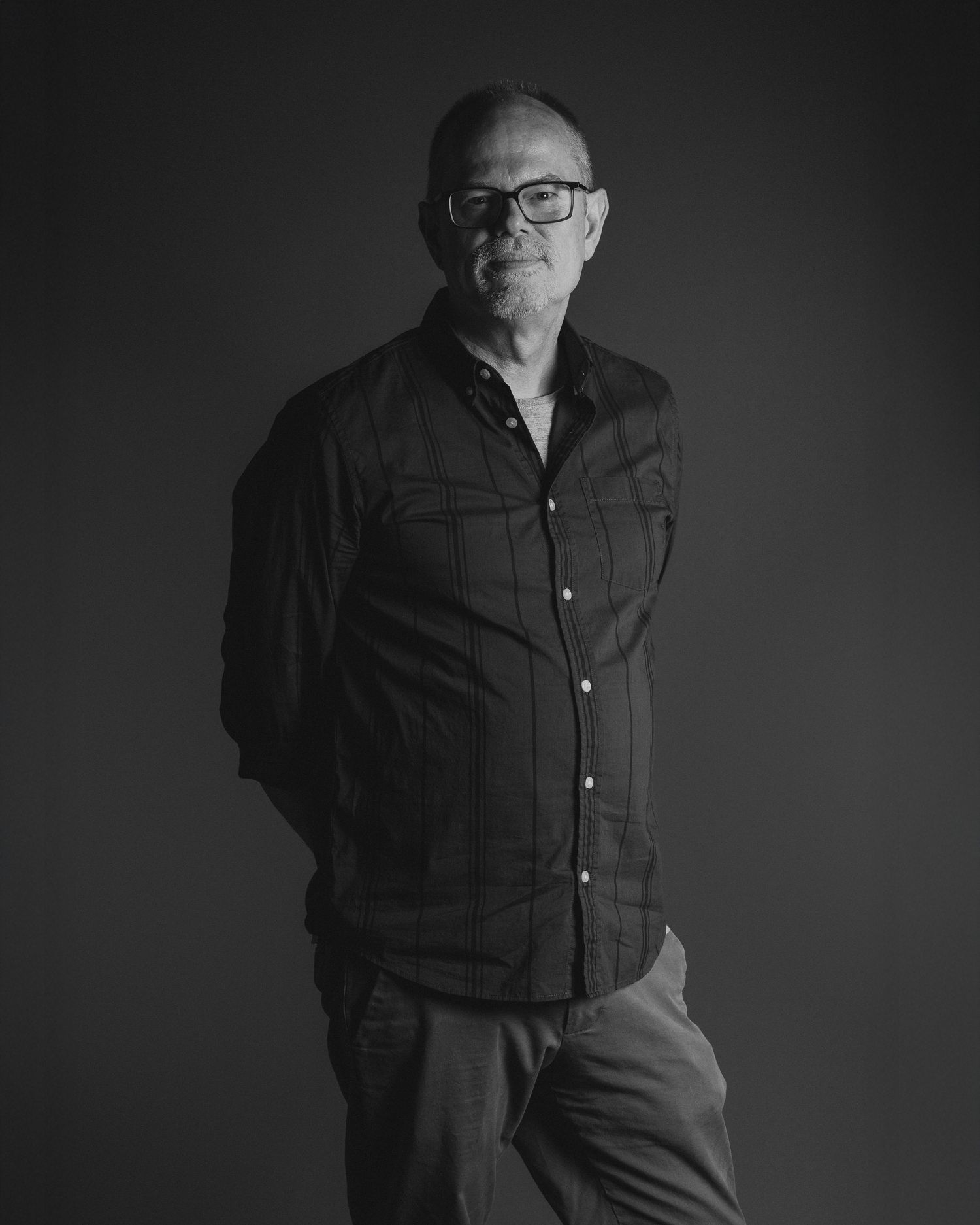 Black and white portrait of a person wearing glasses and casual attire standing against a dark background.